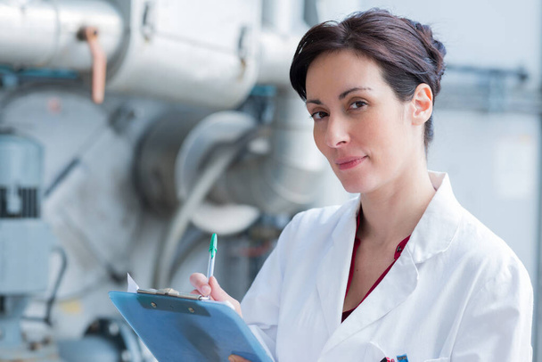woman wearing labcoat and holding clipboard in industrial setting - Foto, afbeelding