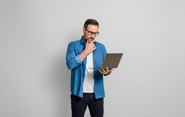 Задумчивый молодой бизнесмен трогает подбородок и работает над ноутбуком изолирован на сером фоне - Фото, изображение