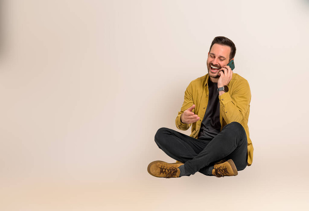 Handsome male manager laughing and communicating over mobile phone while sitting on background. Young professional dressed in shirt talking cheerfully on cellphone - Zdjęcie, obraz
