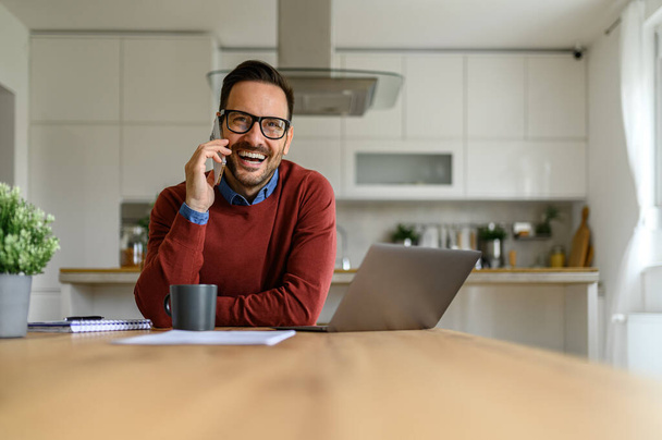 Porträt eines glücklichen männlichen Unternehmers, der geschäftlich telefoniert und im Homeoffice am Laptop arbeitet - Foto, Bild