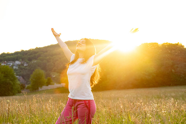 Eine Frau steht mit erhobenen Händen in der untergehenden Sonne im Sommer auf einem Feld in der Natur. - Foto, Bild
