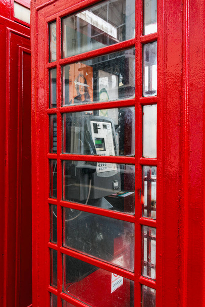 Słynna brytyjska czerwona budka telefoniczna z automatem w środku i szklanymi oknami. Biała klawiatura i ekran. Brudne szkło, naklejka. Miejska scowość z budynkiem w tle. - Zdjęcie, obraz