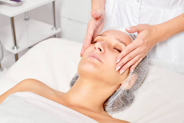Kosmetyk masuje kobiecą skórę twarzy w celu odmłodzenia, zabieg kosmetyczny przeciw starzeniu się skóry w salonie SPA. Kosmetolog robi masaż twarzy kremem kosmetycznym dla pięknej kobiecej twarzy - Zdjęcie, obraz