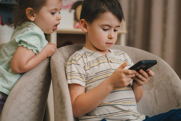 Children with mobile phone playing video games together at home. Game and learning app, study and gadget addiction at spare time at home - Fotografie, Obrázek