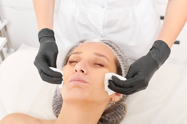 美容用スパサロンで女性の顔の皮膚からフェイシャルクリームマスクを取り除く化粧品専門医. 魔法使いの手は顔のマスクを取除くためのナプキンを握ります - 写真・画像