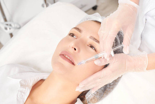 Cosmetologist makes fillers injection for lips augmentation and volume, non surgical cosmetic procedure in beauty salon. Beautician specialist hands in gloves makes lips injection with syringe - Photo, Image