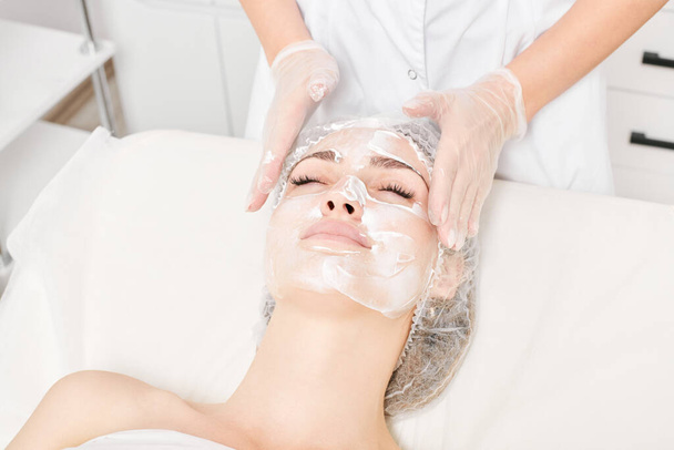 Il cosmetologo massaggia la maschera alla panna in pelle di faccia di donna per ringiovanimento, procedura cosmetica di cura della pelle di invecchiamento in salone di bellezza termale. Estetista mani in guanti sfregamento crema in faccia femminile - Foto, immagini