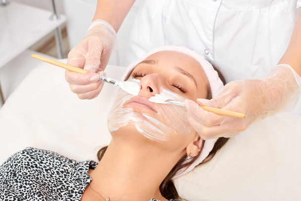 Cosmetologist applying cosmetic cream mask on woman face for rejuvenation face skin, moisturizing cosmetic procedure in spa salon. Beautician in gloves applying cosmetic cream mask with two brushes - Photo, Image