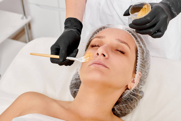 美容師は,顔の保湿,美容スパサロンでの老化防止化粧品手順のために女性の顔に蜂蜜マスクを適用します. ブラックグローブのビーガーは蜂蜜のマスクを適用するための化粧ブラシを保持します - 写真・画像