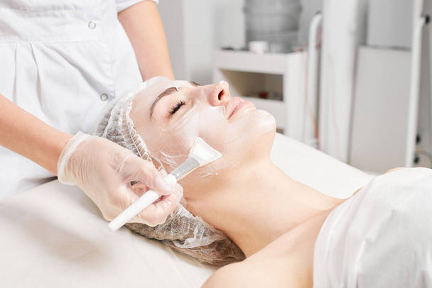 Kozmetológus alkalmazza krém maszk a nő arcát fiatalító arc bőr, anti aging kozmetikai eljárás szépségszalonban. Kozmetikus kéz kesztyűben tartja kozmetikai kefe és alkalmazása kozmetikai maszk - Fotó, kép