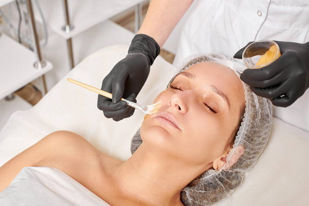 Cosmetologist applies honey mask on woman face for moisturize face skin, anti aging cosmetic procedure in beauty spa salon. Beautician in black gloves holds cosmetic brush for applying honey mask - Photo, Image