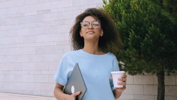 Elegante ragazza afroamericana con i capelli ricci, donna freelance di razza mista, studentessa, con gli occhiali, in camicia blu, tenendo il computer portatile, camminando all'aperto, guardandosi intorno, sorridendo felicemente - Filmati, video