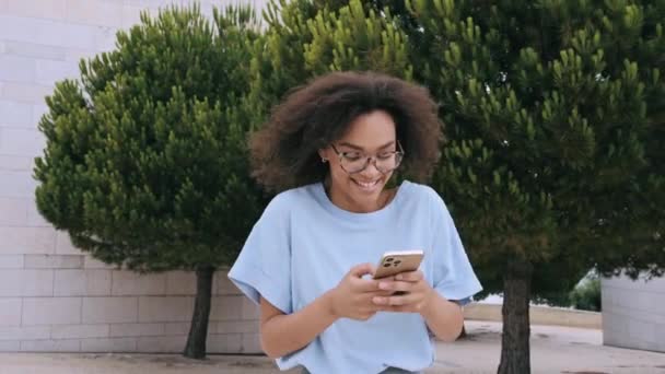 Positivo satisfeito afro-americano encaracolado jovem mulher, elegantemente vestida, usando seu smartphone enquanto caminha ao ar livre, conversando com amigos em redes sociais, responde e-mail, sorrindo  - Filmagem, Vídeo