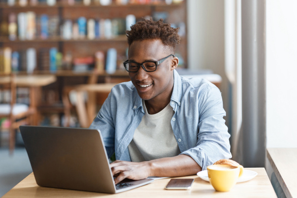 Positive handsome millennial black guy wearing casual outfit and eyewear sitting at desk in front of computer laptop, freelancer working online at cafe, drinking coffee, copy space - Photo, Image