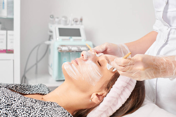 Kosmetolog stosujący kosmetyczną maskę kremową na twarzy kobiety do odmładzania skóry twarzy, nawilżający zabieg kosmetyczny w salonie spa. Kosmetyczka w rękawiczkach z maską kosmetyczną z dwoma pędzlami - Zdjęcie, obraz