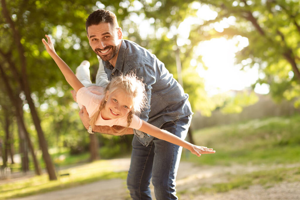 Ενθουσιασμένο κοριτσάκι να παίζει με τον ευτυχισμένο μεσήλικα πατέρα της στο πάρκο, παιδί να τεντώνει τα χέρια, να προσποιείται ότι πετάει. Ο μπαμπάς κρατάει την κόρη του, περνάει καλά μαζί. - Φωτογραφία, εικόνα