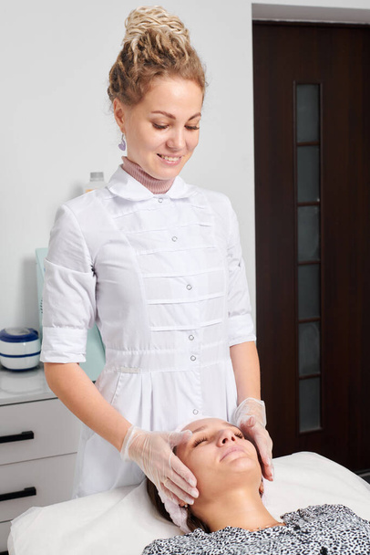 Beautician in gloves massages female face skin for rejuvenation, face lymphatic drainage, cream skincare cosmetic procedure in beauty spa salon. Cosmetologist hands smoothing face skin for procedure - Photo, Image