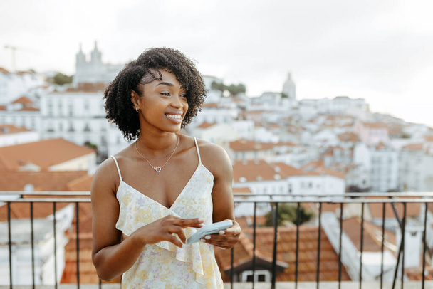 Smiling young african american lady in dress walks in city, looks at empty place, enjoys travel with smartphone, outdoor. Chat, app, blog and map for vacation trip, weekend and holiday alone - Foto, Imagen