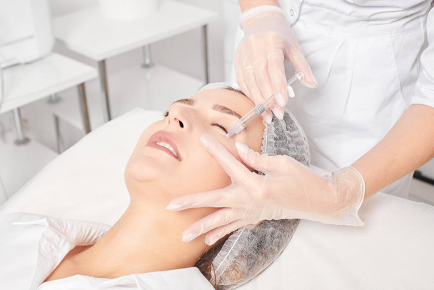 Cosmetologist makes rejuvenation injection in woman face skin, anti aging revitalization cosmetic procedure in beauty salon. Beautician hands in gloves makes facial acid injection treatment - Photo, Image