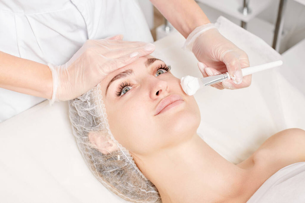 Kozmetikus tesz krém maszk a nő arcát fiatalító arc bőr, anti-aging kozmetikai eljárás szépségszalonban. Kozmetológus kéz kesztyűben tartja kozmetikai kefe és alkalmazza frissítő maszk - Fotó, kép