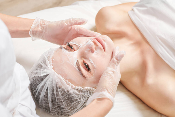 Beautician massages cream mask into female face skin for rejuvenation, moisturizing skincare cosmetic procedure in beauty spa salon. Cosmetologist hands in gloves rubbing cream in woman face - Photo, Image