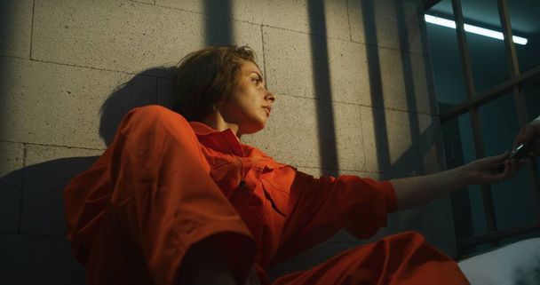 Gefängniswärter gibt Telefon an weibliche Gefangene. Kriminelle Frau in orangefarbener Uniform ruft an, sitzt auf dem Bett in der Gefängniszelle. Frau verbüßt Haftstrafe im Gefängnis Haftanstalt oder Justizvollzugsanstalt. - Foto, Bild