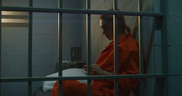 Egy női rab okostelefont használ a börtönben. Narancssárga egyenruhás bűnöző nő telefonál, az ágyon ül a cellában. Börtönben ül. Fogolytábor vagy büntetés-végrehajtási intézet. - Fotó, kép