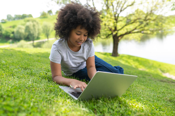 Charming little girl with curly hair using laptop in summer garden having video call watching movie online lesson distance education. - Foto, Imagem