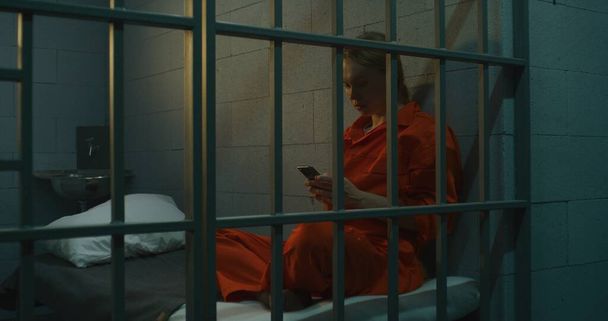Więźniarka używająca smartfona w więzieniu. Kobieta w pomarańczowym mundurze dzwoni, siedzi w więziennej celi. Odsiaduje wyrok więzienia w więzieniu. Centrum zatrzymań lub zakład karny. - Zdjęcie, obraz
