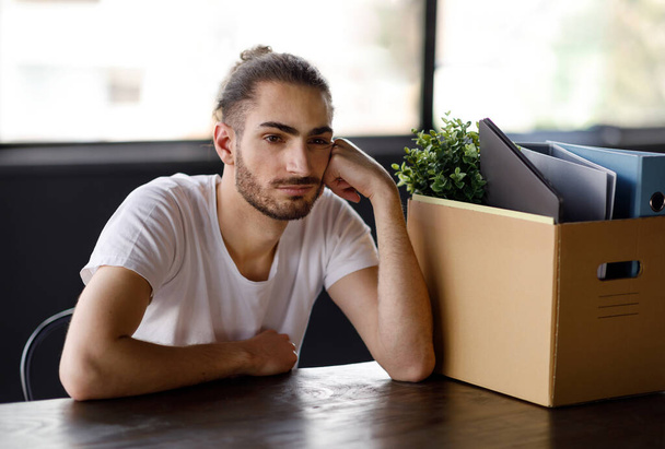 Απόλυση, οικονομική κρίση. Δυστυχισμένος Ισπανόφωνος νεαρός άνδρας κάθεται αναστατωμένος κοντά σε προσωπικά αντικείμενα στο κουτί αφού απολύθηκε στο εσωτερικό του γραφείου. Άνεργος που αισθάνεται κατάθλιψη μετά την απώλεια της δουλειάς του - Φωτογραφία, εικόνα