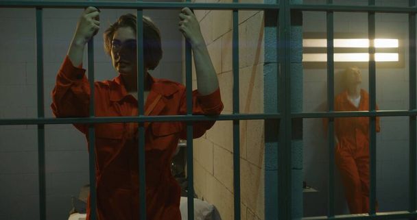Een vrouwelijke gevangene in oranje uniform staat achter metalen tralies, een andere zit op het bed in de cel. Vrouwen zitten gevangenisstraf uit voor misdaden in de gevangenis. Depressieve gevangenen in detentiecentrum. - Foto, afbeelding