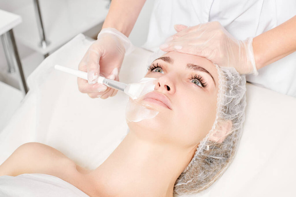 Kozmetológus alkalmazza krém maszk a nő arcát fiatalító arc bőr, anti aging kozmetikai eljárás szépségszalonban. Kozmetikus kéz kesztyűben tartja kozmetikai kefe és alkalmazása kozmetikai maszk - Fotó, kép