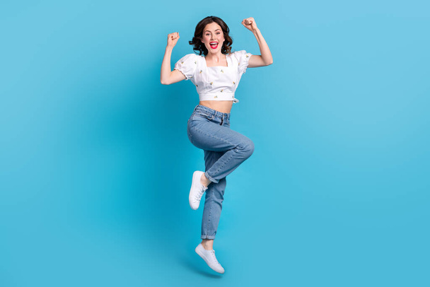 Full length photo of cute lucky lady wear white blouse jumping high rising fists isolated blue color background. - Фото, зображення