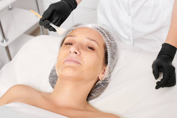 Beautician εφαρμόζει μάσκα μελιού στο πρόσωπο της γυναίκας για ενυδάτωση του δέρματος του προσώπου, αντιγηραντική καλλυντική διαδικασία στο σαλόνι ομορφιάς spa. Cosmetologist σε μαύρα γάντια κατέχει καλλυντική βούρτσα για την εφαρμογή μάσκα μελιού - Φωτογραφία, εικόνα