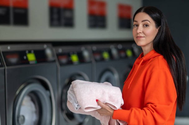 Laundry service. Brunette adult woman wearing orange hoodie holding washed towels in public laundry. - 写真・画像