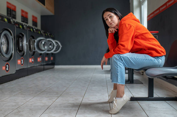 Tired bored woman wearing orange hoodie sitting in automatic laundry room while waiting for the laundry to be done. - Fotó, kép