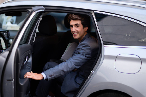 Wesoły, przystojny młodzieniec w garniturze, przedsiębiorca wsiadający do szarego auta. Milenijny biznesmen lub menedżer zamykający drzwi samochodu i uśmiechający się do kamery. Koncepcja podróży służbowej - Zdjęcie, obraz