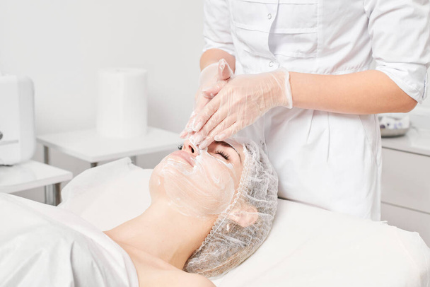 Cosmetologist μασάζ κρέμα μάσκα προσώπου σε γυναίκα δέρμα για αναζωογόνηση, αντι γήρανση φροντίδα του δέρματος καλλυντική διαδικασία στο σαλόνι ομορφιάς spa. Όμορφα χέρια σε γάντια τρίψιμο κρέμα στο γυναικείο πρόσωπο - Φωτογραφία, εικόνα