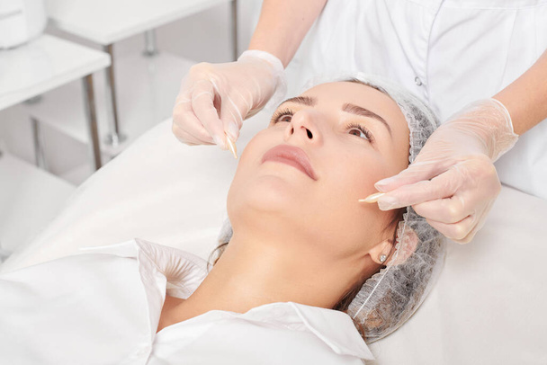 Kosmetolog aplikuje kapsle oleje na obličej ženy pro omlazení, proti stárnutí kosmetické procedury v kosmetickém salonu. Kosmetik ruce v rukavicích drží olejové kapsle pro výživu - Fotografie, Obrázek