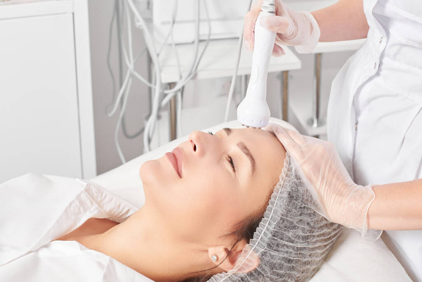Cosmetologist makes RF lifting for rejuvenation woman face, anti aging cosmetic procedure in beauty spa salon. Beautician makes radio frequency treatment for collagen increasing on female face skin - Photo, Image