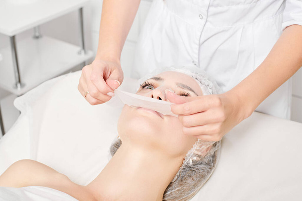 Beautician αφαιρώντας μάσκα σεντόνι στο πρόσωπο της γυναίκας για ενυδατική επιδερμίδα προσώπου, αντιγηραντική καλλυντική διαδικασία στο σαλόνι ομορφιάς spa. Cosmetologist αφαίρεση ενυδατική μάσκα φύλλο στο γυναικείο πρόσωπο - Φωτογραφία, εικόνα