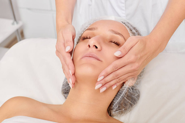 Косметологічний масаж жінки обличчям до шкіри обличчя для омолодження, анти-старіння косметичної процедури шкіри в салоні краси. Косметолог робить масаж обличчя косметичним кремом для красивого жіночого обличчя - Фото, зображення