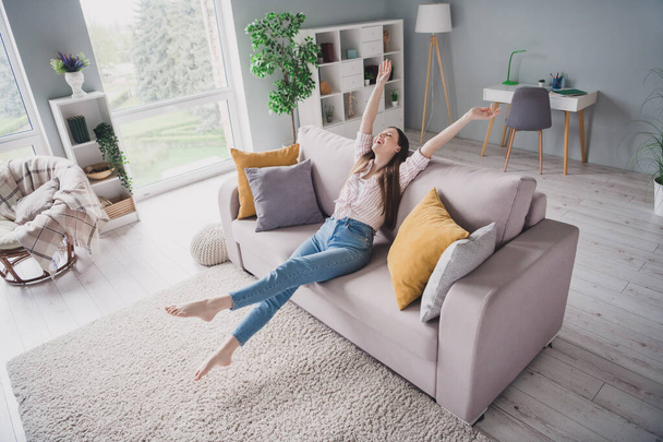 Photo of shiny carefree young girl wear striped shirt stretching rising arms hands sitting sofa indoors apartment room. - Foto, Imagem