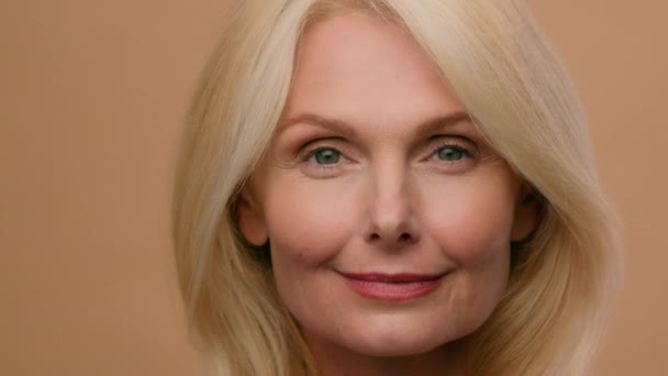 Headshot mature Caucasian 50s woman middle-aged female beautiful model looking at camera anti wrinkle age cosmetic natural makeup beauty skincare face lifting healthy toothy smile on beige background - Záběry, video