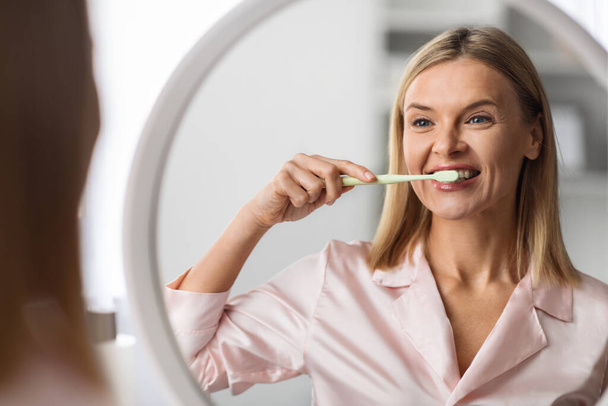 Belle femme mûre se brossant les dents avec une brosse à dents près du miroir, heureuse femme du Moyen Âge souriant à son reflet, rendant l'hygiène du matin à la maison, faisant des soins buccodentaires, se concentrant sélectivement - Photo, image
