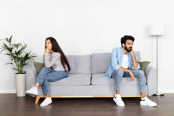 Crisis in relationships, marriage, breakup, divorce. Upset frustrated young eastern couple sitting on couch back to back, dont talk to each other after fight, white wall background, home interior - 写真・画像