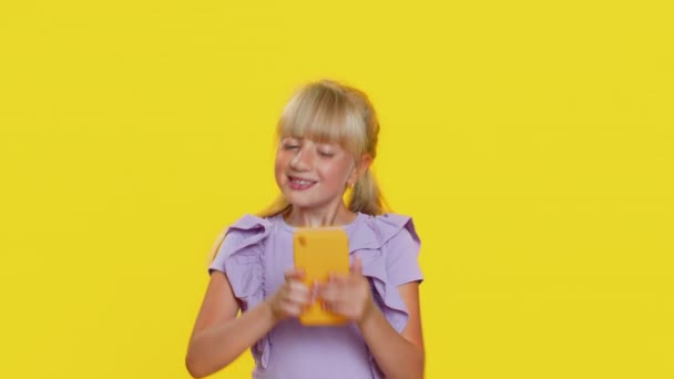 Young preteen child girl kid use mobile smartphone typing browsing say Wow yes found out great big win good news celebrate doing winner gesture. Little cute children on studio yellow background - Footage, Video