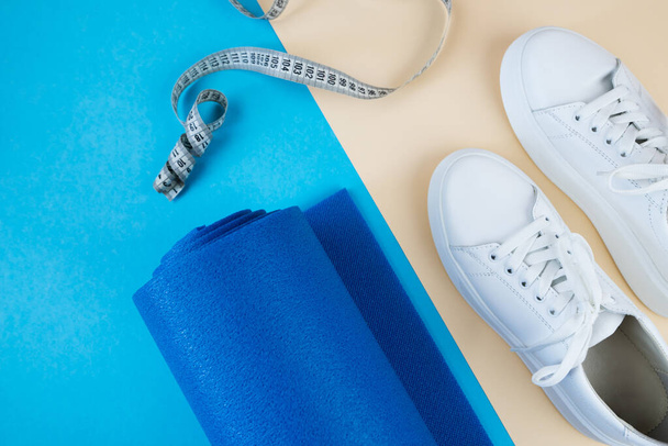 The concept of sports accessories. A photo of white sneakers, blue dumbbells, a blue exercise mat and other sports equipment against a pastel beige background. View from above. - Photo, image