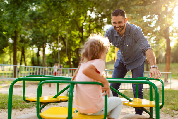 Child girl having fun with her father on outdoor playground, dad riding daughter on carousel, spending active leisure time together. Happy parenthood and childhood - Photo, Image