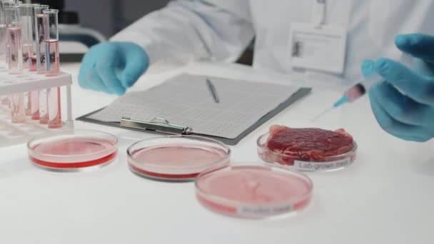 Close-up of gloved microbiologist injecting plant-based serum in in-vitro meat sample in petri dish while sitting by workplace in clinical laboratory - Footage, Video